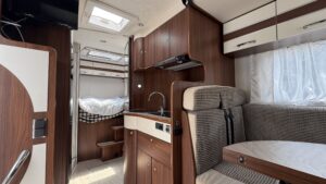 Interior de autocaravana con cocina compacta, salón comedor, armarios superiores y cama trasera elevada con acceso mediante escalones.