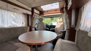 Interior amplio y luminoso de autocaravana con salón comedor en forma de U, mesa central extensible, asientos delanteros giratorios y claraboya panorámica.