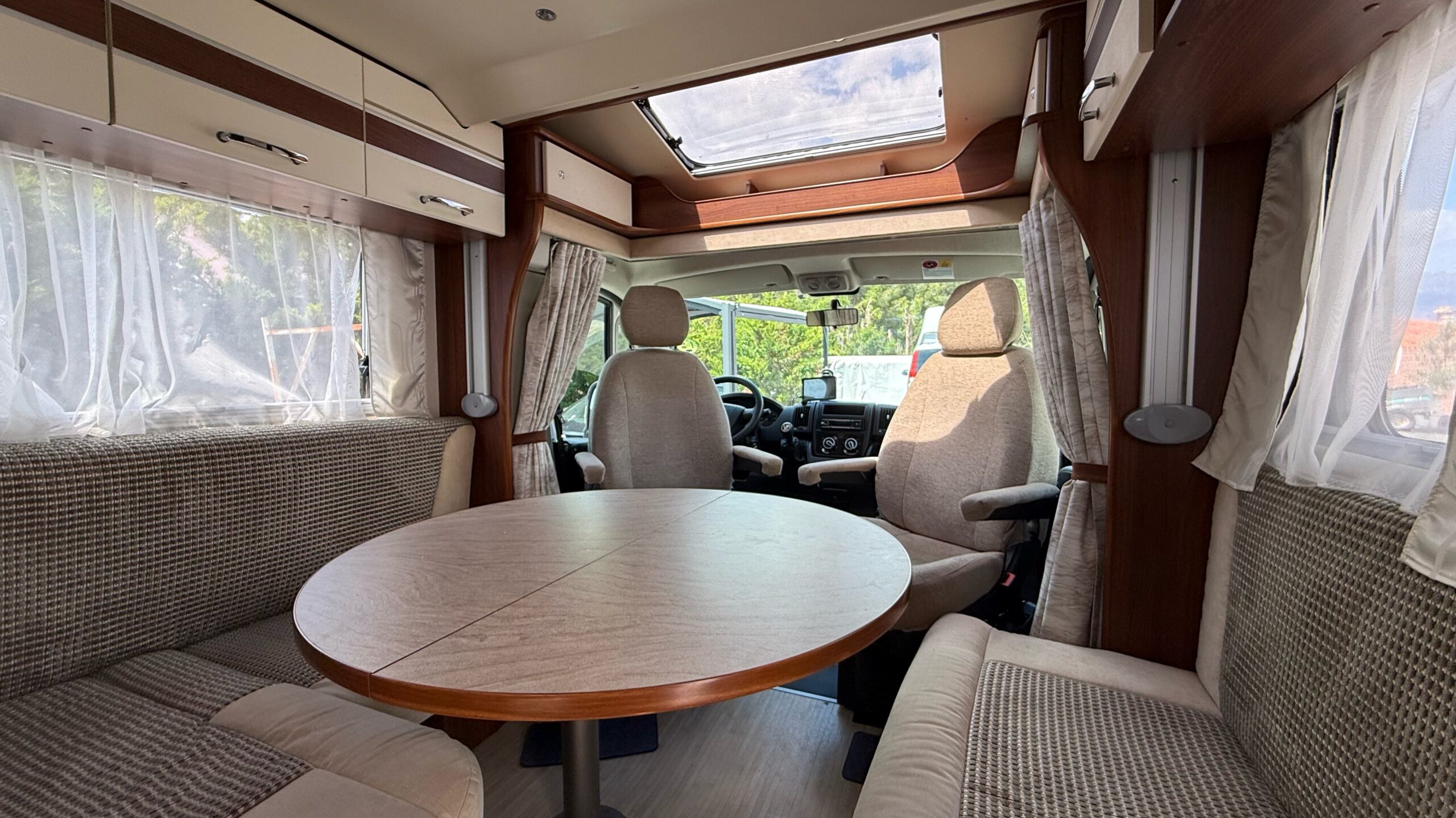Interior amplio y luminoso de autocaravana con salón comedor en forma de U, mesa central extensible, asientos delanteros giratorios y claraboya panorámica.